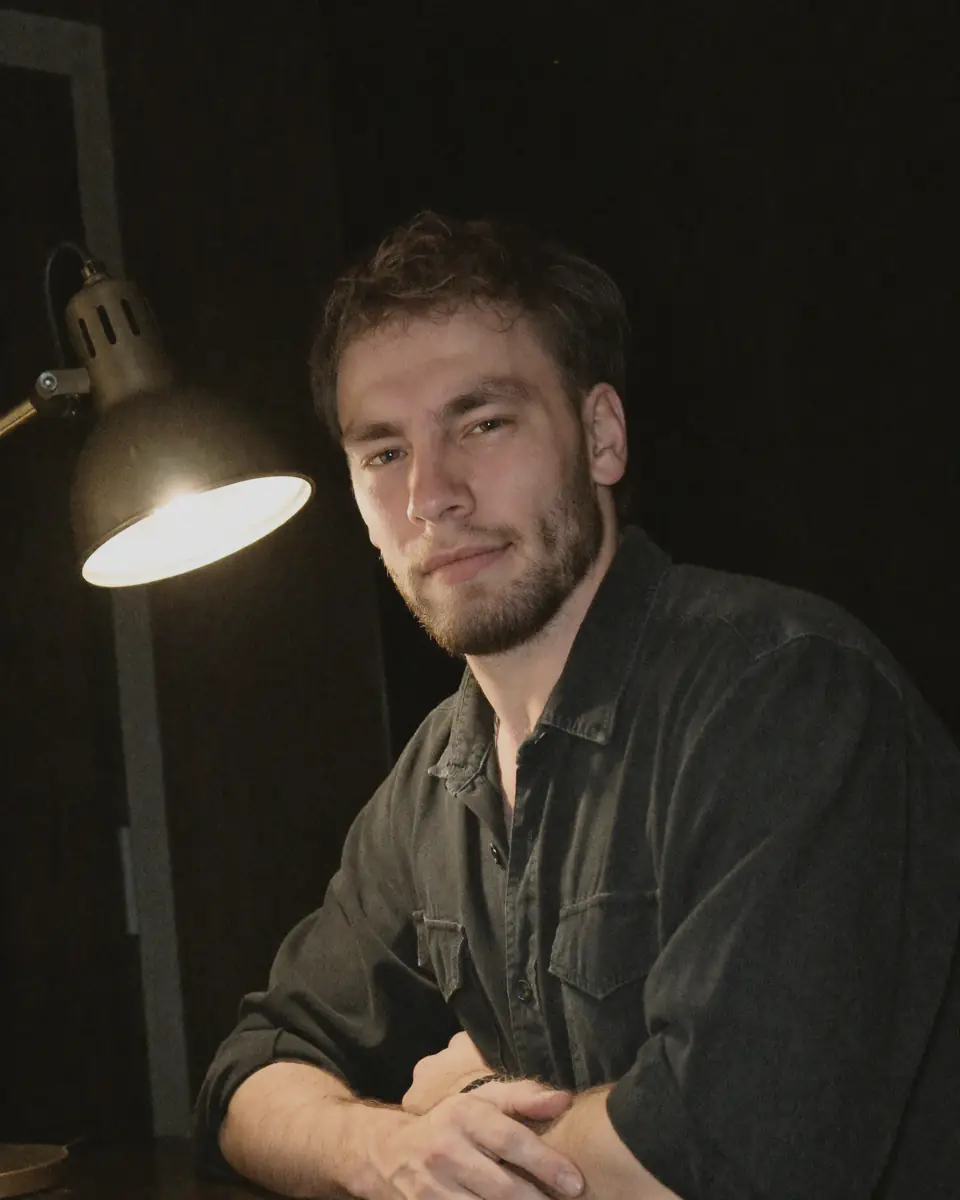 Jules sitzt mit verschränkten Armen an seinem Arbeitstisch unter einer Lampe und lächelt in die Kamera.